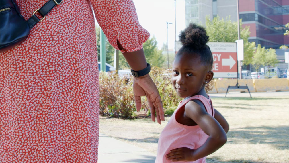 Preschool age girl in a pink tank top stands outdoors with her hands on hips, next to a woman’s outstretched hand