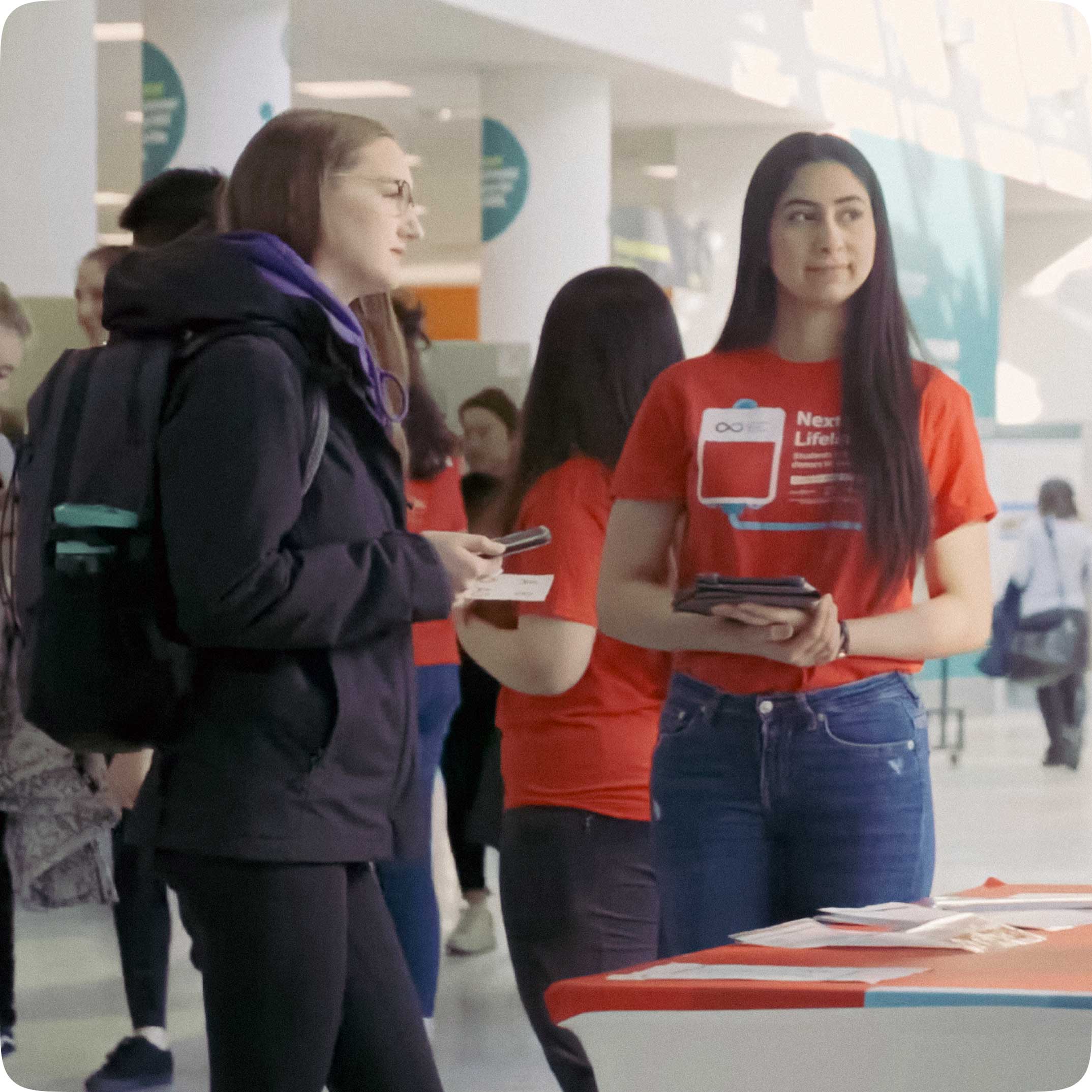 A person with a backpack and a Canadian Blood Services staff member standing in front of a table with collateral, looking off to the side together, with a busy background of people and tables.
