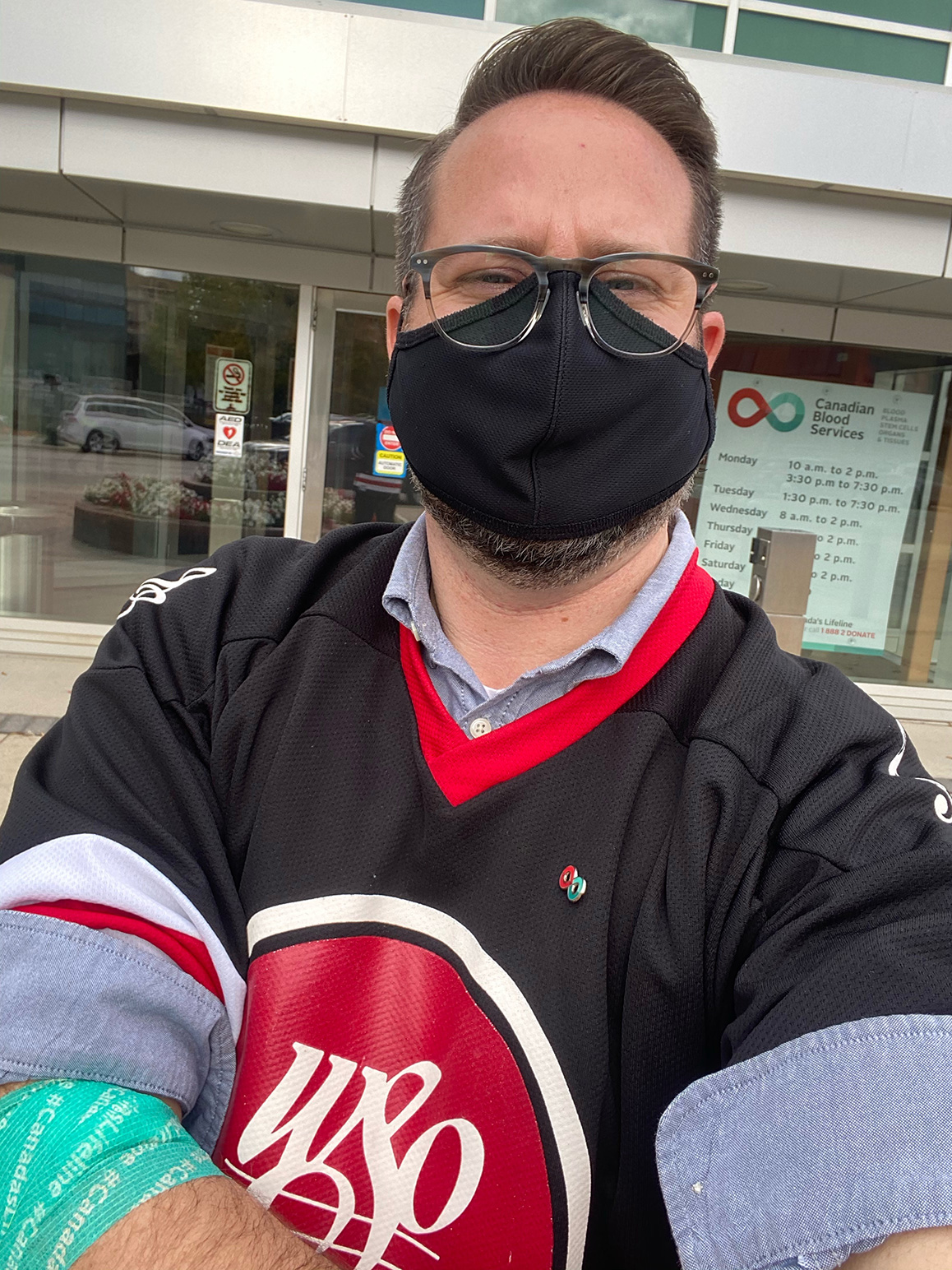 Image of blood donor Brent Johnson wearing a mask and a black jersey with blue shirt standing outside of a blood donor.