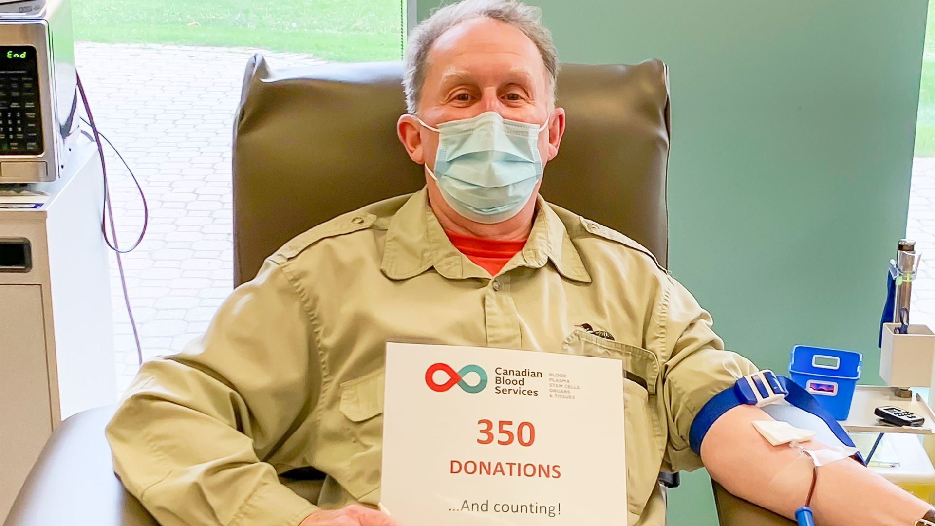 A man is wearing a mask while seated in a reclining chair and holding a sign that reads 350 donations and counting! as blood is drawn from his arm.