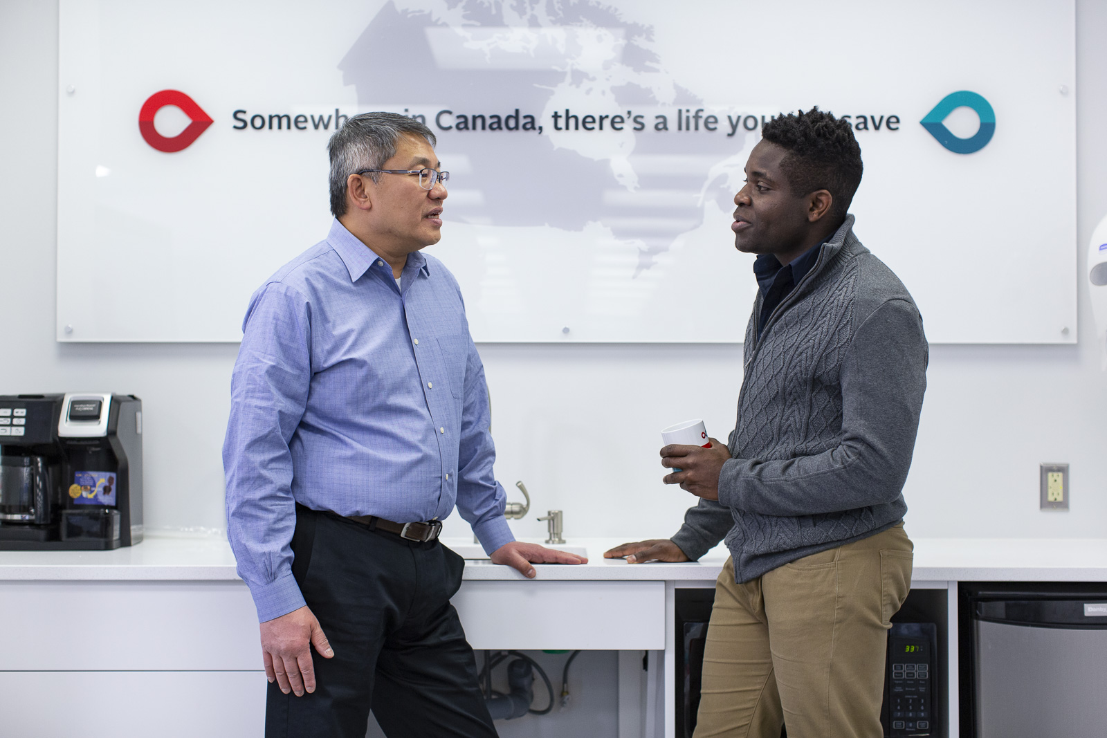 An Asian employee and a Black employee in conversation. Behind them is a map of Canada with the words Somewhere in Canada there’s a life you can save.