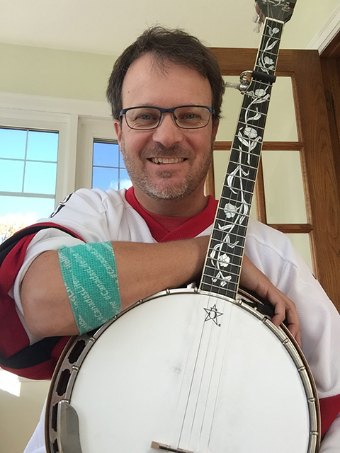 Image of Gregory Hay holding a banjo and wearing a white shirt with a blood donor badge