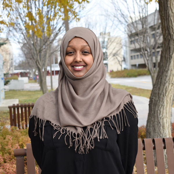 Photo of Razia Seman standing in front of a bench and a tree out a neighbourhood park wearing a hijab