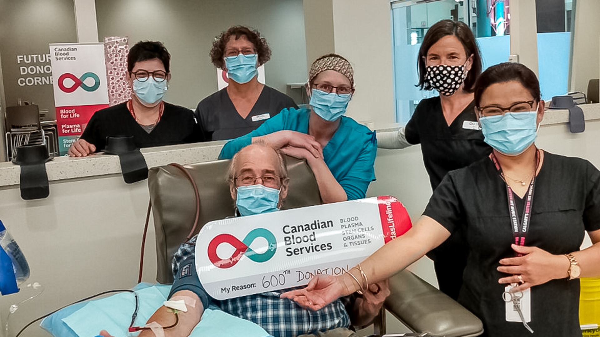 Image of Ian Harrop holding up the sign of My Reason for his 600th donation with the nurses