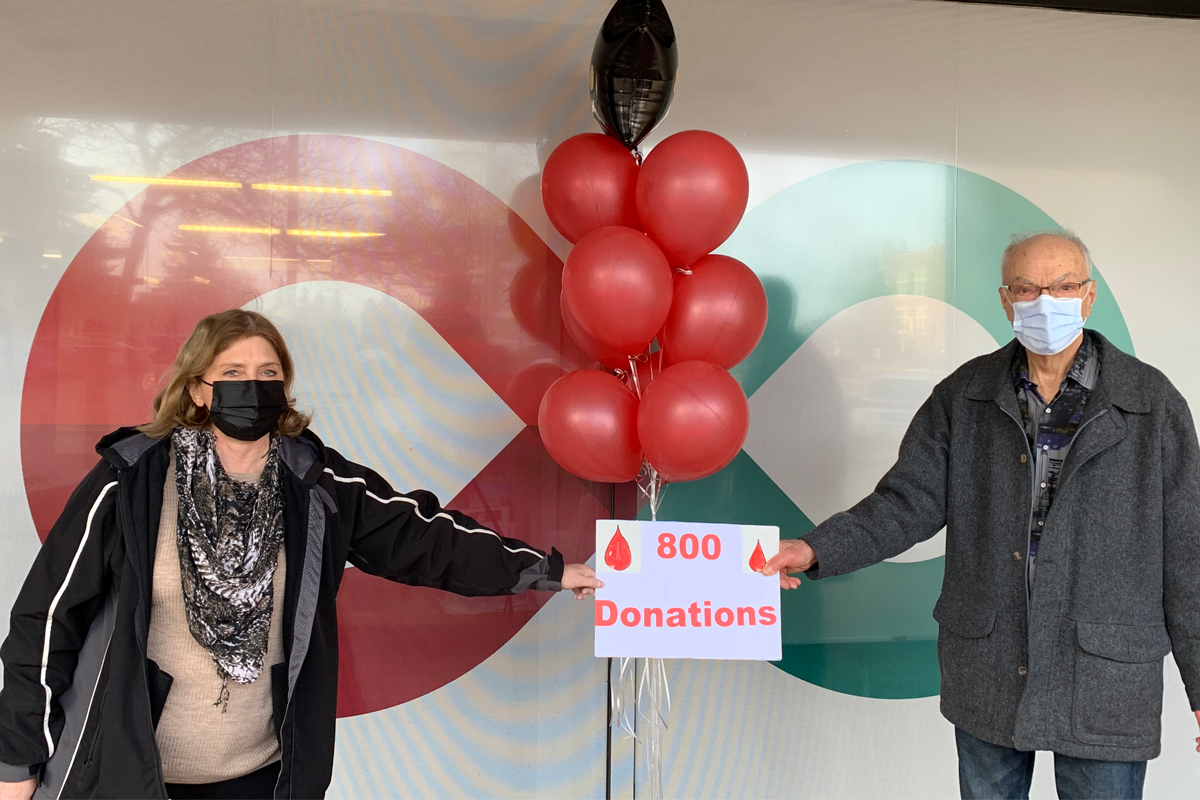 Allan and Lorraine standing outside a donor centre holding a sign that says 800 donations for plasma.