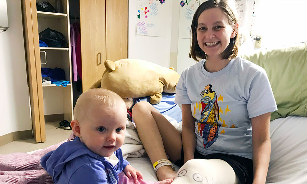 Young woman whose left leg has been amputated and bandaged smiling on a bed with a young baby sitting next to her
