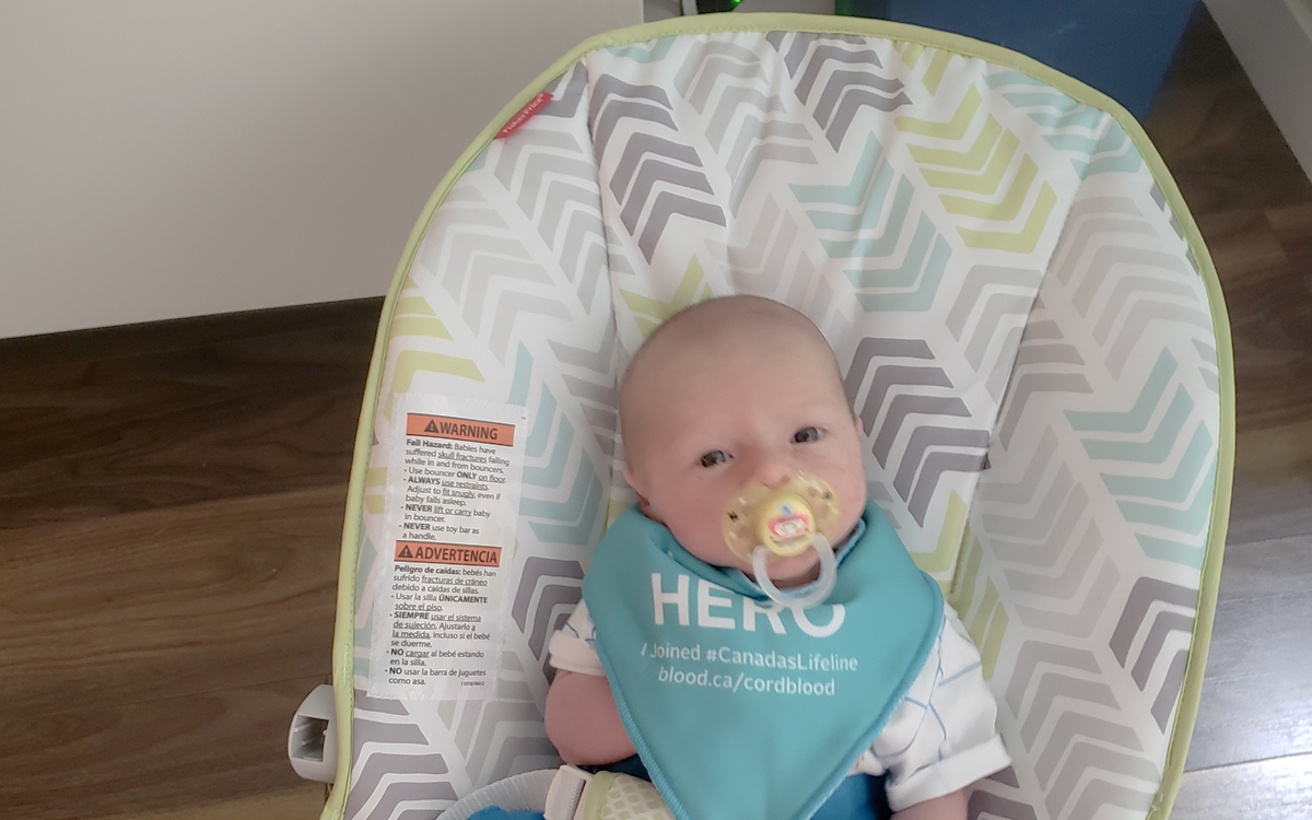 one-month-old baby in a rocker chair with a soother in its mouth and wearing a bib with ‘hero’ written across it
