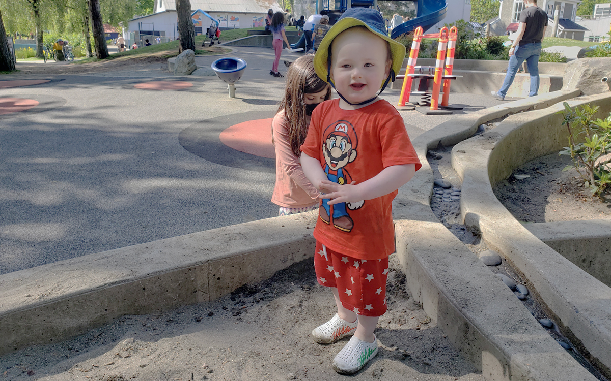 two-year-old boy stands in the corner of a sandbox in a play area with children in the background