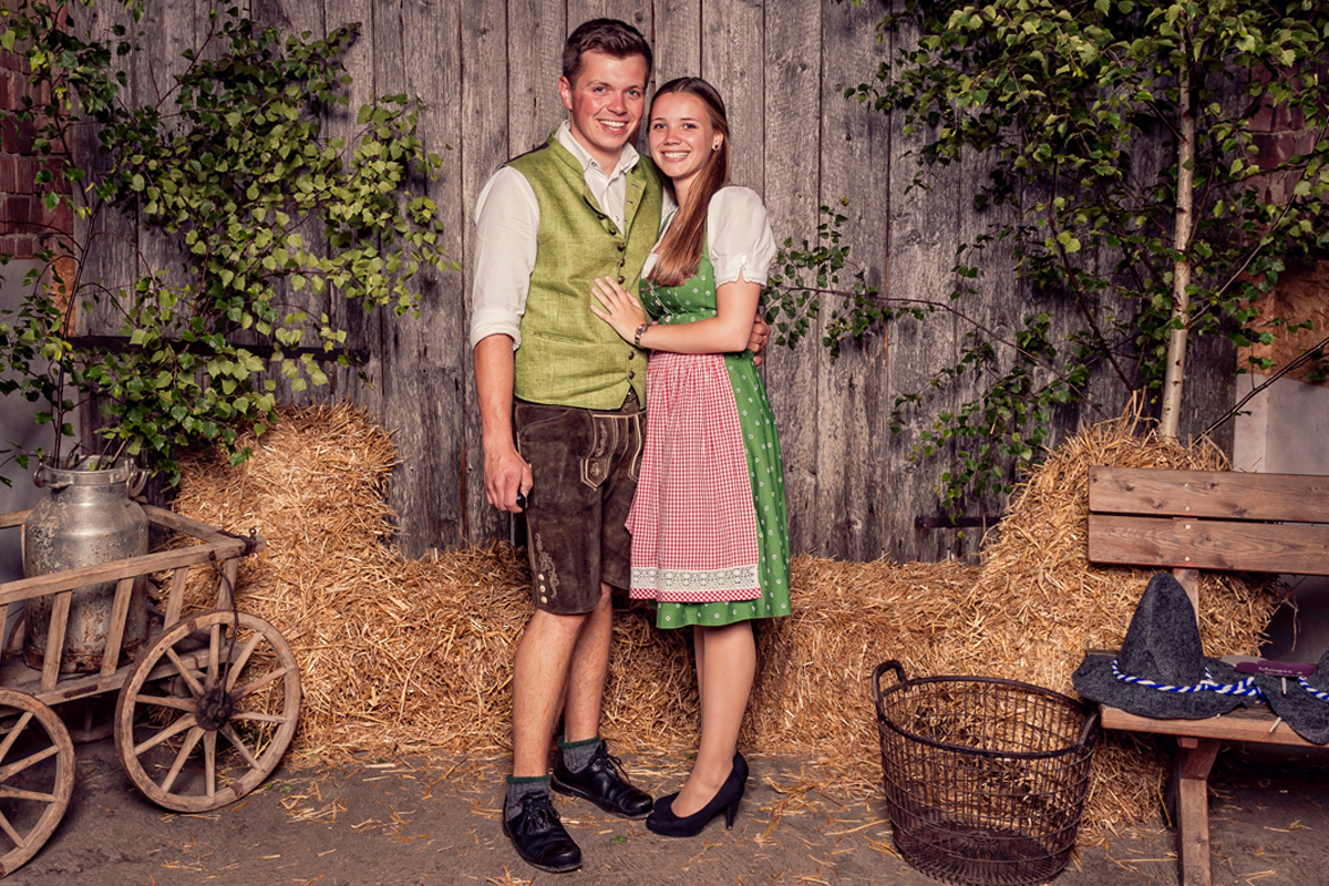 A young couple hold each other outside a house, with loose hay behind them and garden items