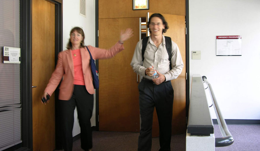 Woman with a salmon coloured blazer with black pants and tall man with long brown hair with a white shirt and black pants entering an office