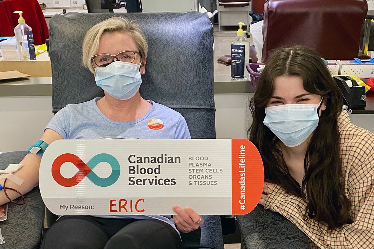 Woman donating blood holds sign that says “Eric” with daughter next to her.