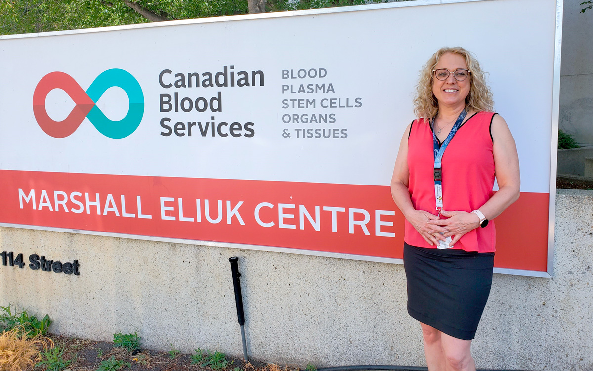 A woman in a coral shirt with long blond hair stands next to the sign for Canadian Blood Services’ Marshall Eliuk Centre.