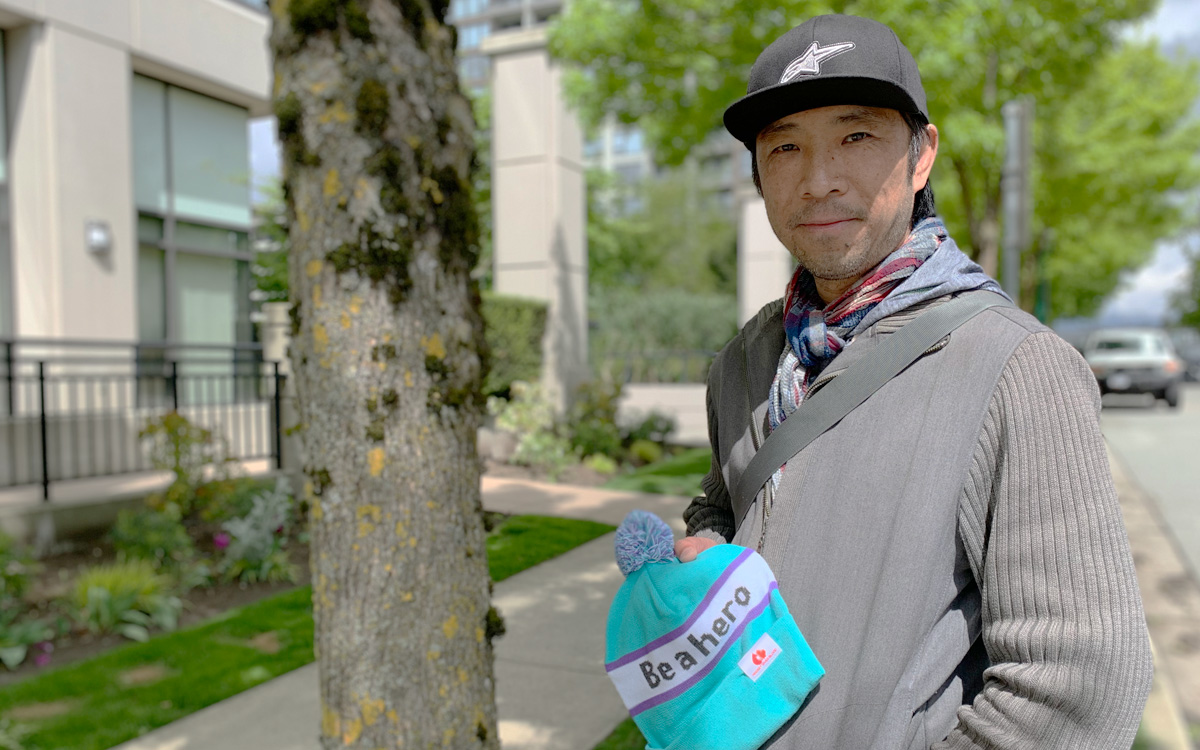 Edmond Chan, in a baseball cap, stands by a tree holding a toque with ‘be a hero’ written across the front