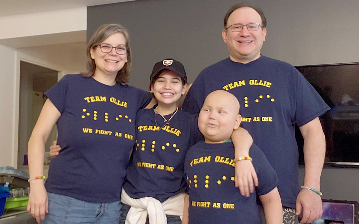 A family of four stands together wearing navy blue and yellow T-shirts that say “Team Ollie: We Fight As One” with the name “Ollie” also spelled in Braille.