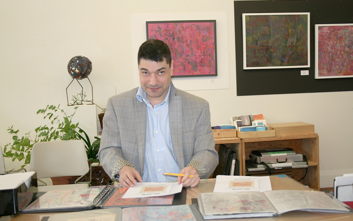 An artist sits at a desk signing autographs with some of his works displayed behind him.