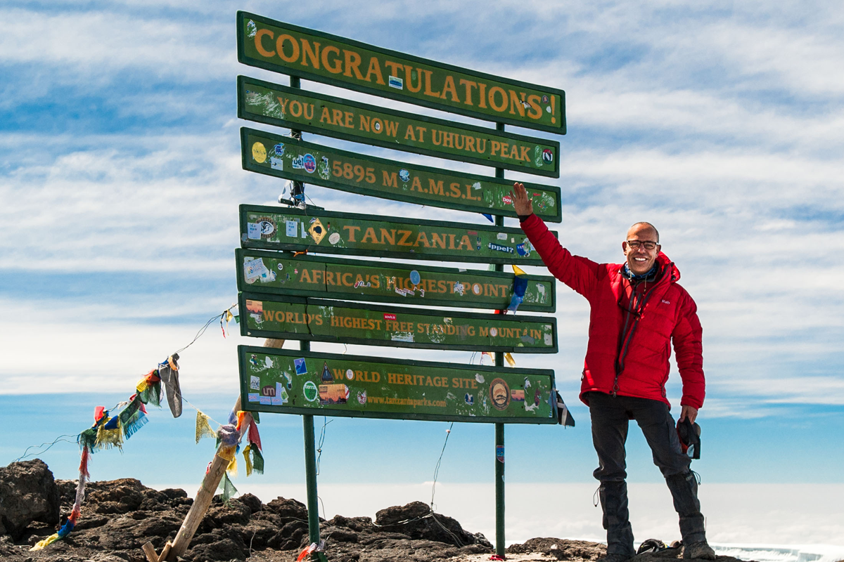 Man in red jacket pointing to sign at Uhuru Peak on Mt. Kilimanjaro, Africa’s highest point.