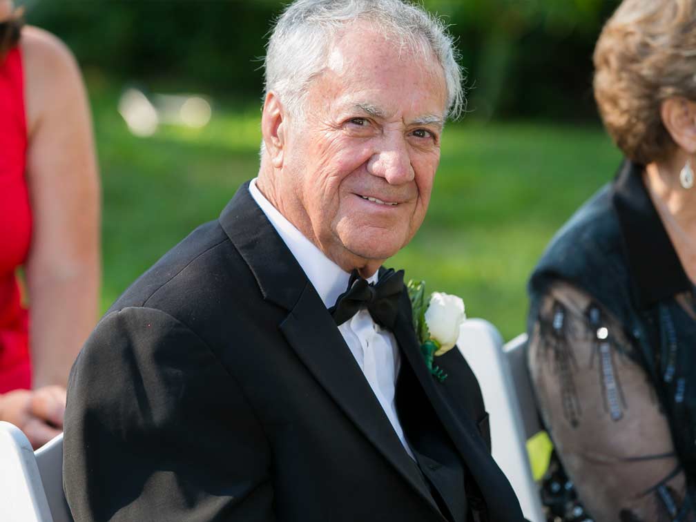 White-haired man in suit with bowtie and corsage sitting at an outdoor wedding