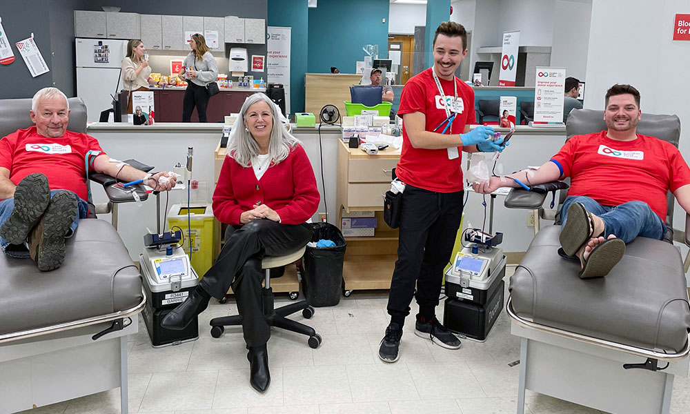 Two men donating blood and an employee standing there wearing red Canadian Blood Services logo shirt.