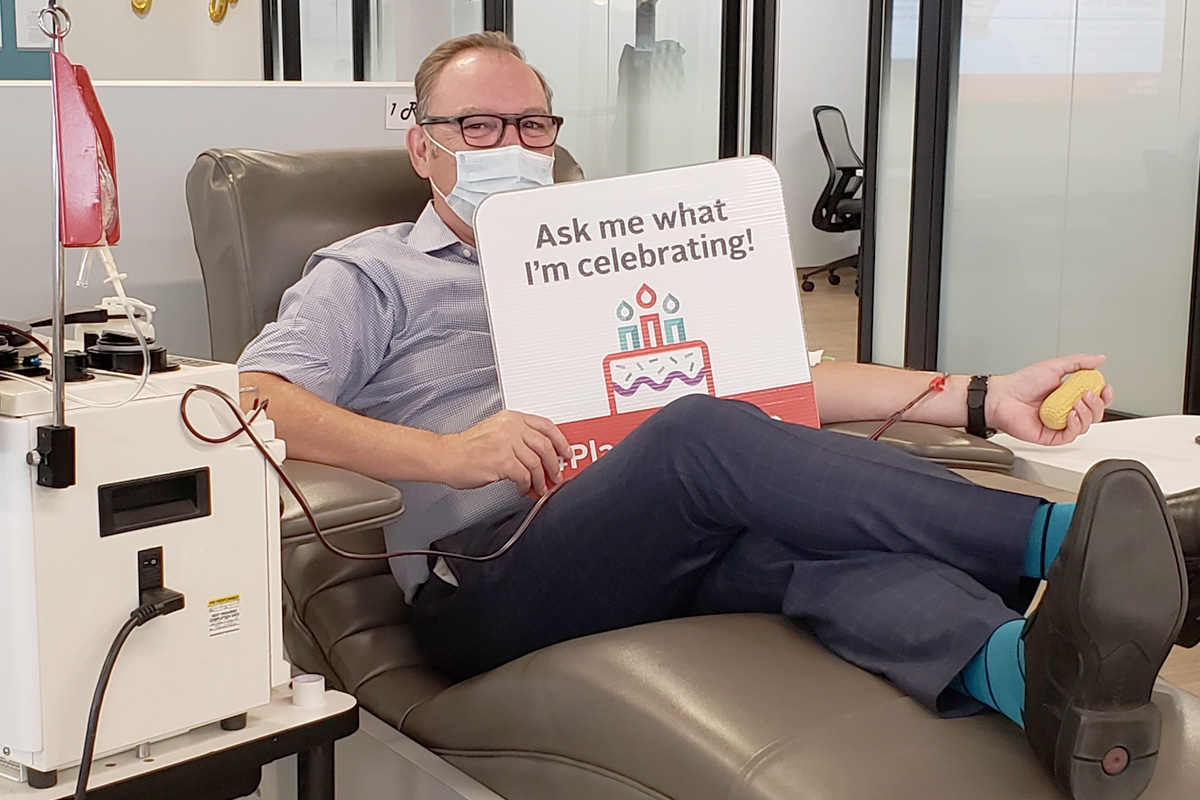 A plasma donor donates plasma while holding a sign that says “Ask me what I’m celebrating!”
