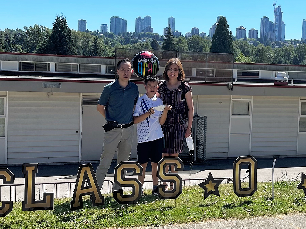 Parents and son outdoors with a balloon behind a sign that says “Class O 2021.”