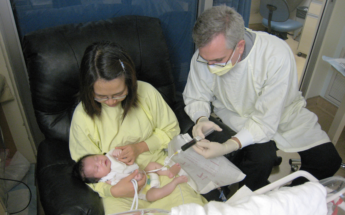 Dressed in hospital gowns, Celia Lau holds her baby on her lap as their doctor administers a chemotherapy drug through a syringe.
