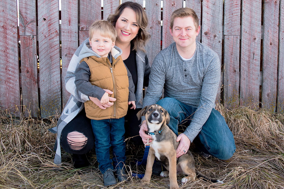A mother and father with their young son and dog in front of a wooden fence.
