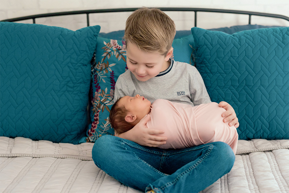 A boy on a bed holds his infant sister who is swaddled in a pink blanket.