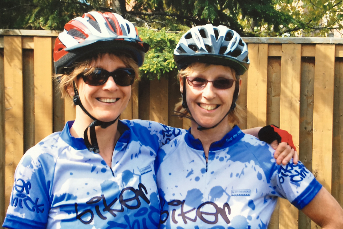 Margaret Lynch and her sister Mary smile in their cycling gear in front of a wooden fence.