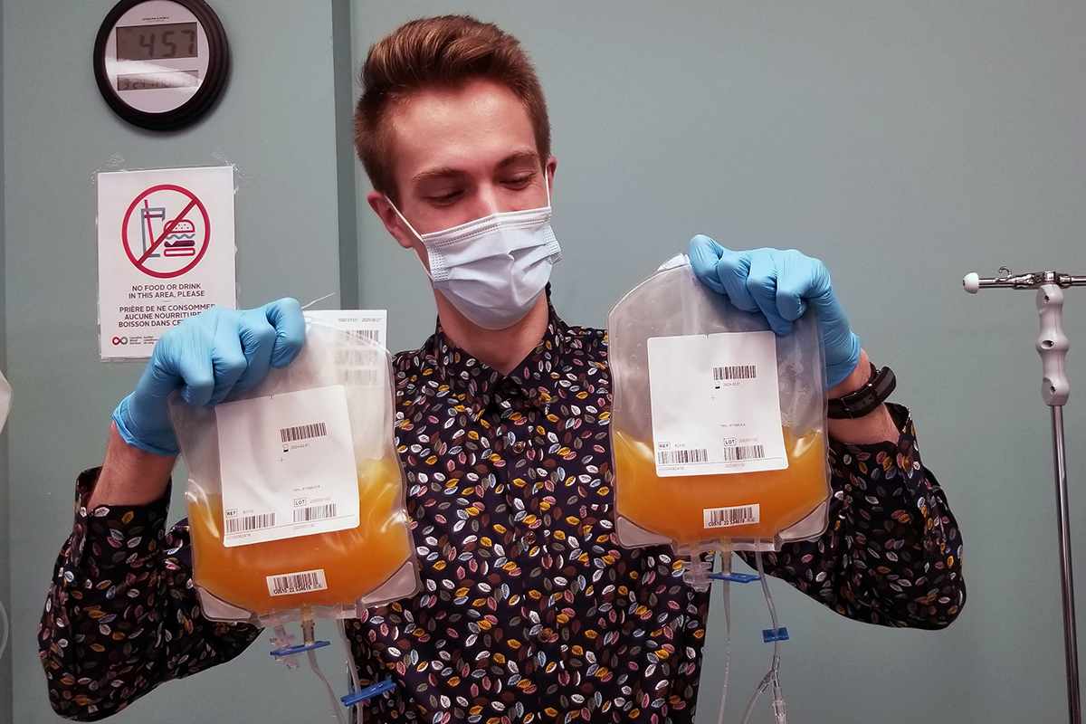 A plasma donor wearing a mask and medical gloves holds a platelet donation in each hand