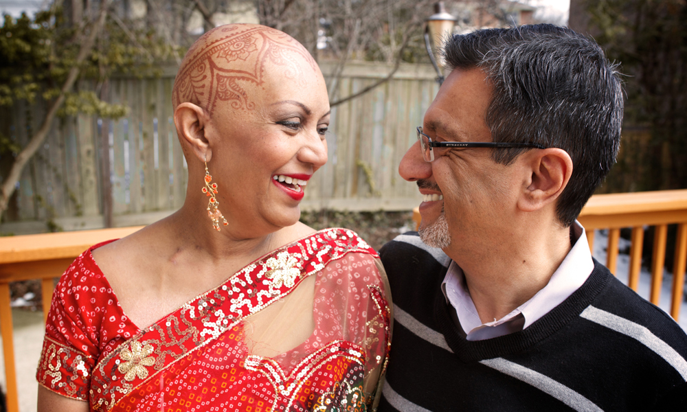 woman who had many blood transfusion procedures during cancer treatment stands next to her husband smiling. She has henna designs decorated on her head.