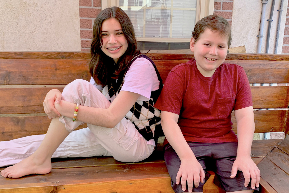 A boy and his sister sit on a bench in their backyard.