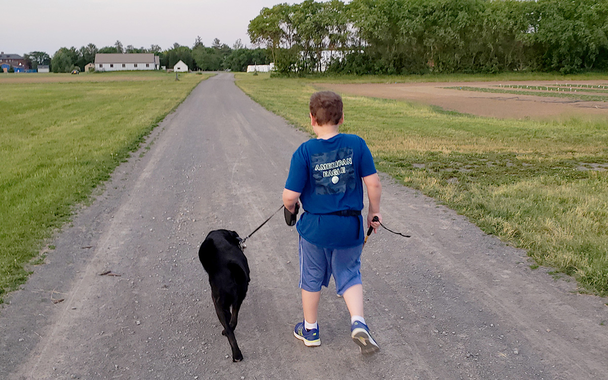 A boy with a cane and black buddy dog on a leash walk down a road together.