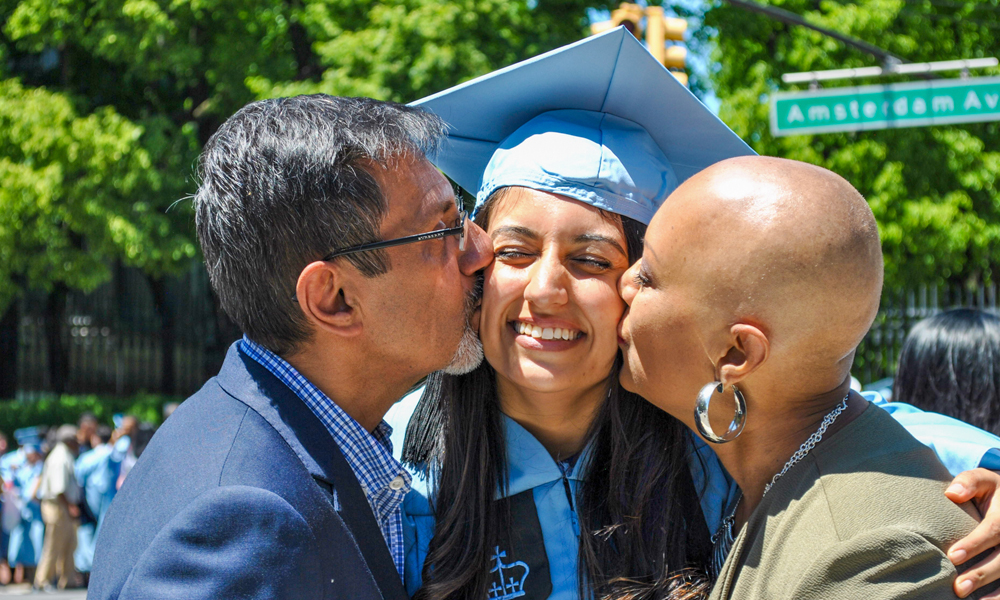 A young woman in a mortarboard receives kisses from her two parents on graduation day in New York City.