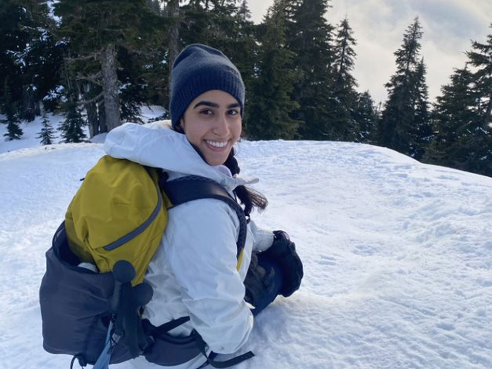 A young Sikh female crouched on top of a snow hill with trees in the background