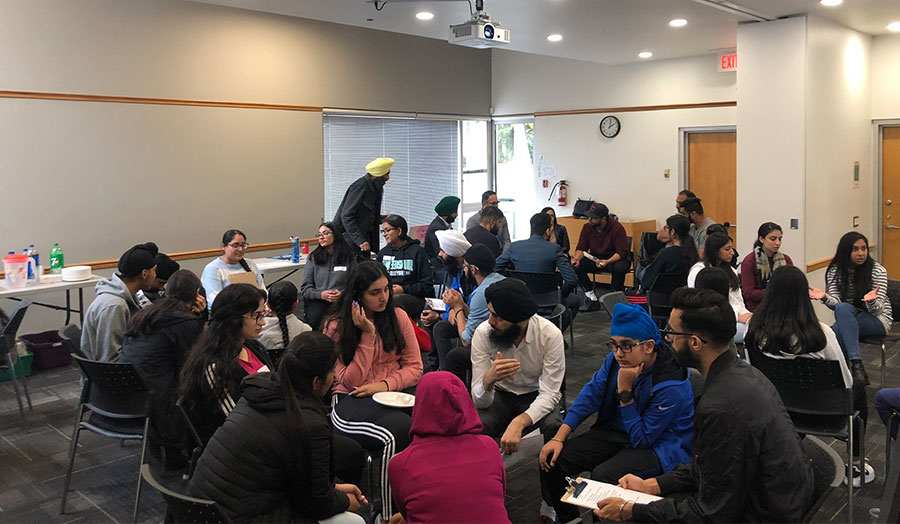 A group of Sikh Nation volunteers sit together in a large room, huddled into small groups. Some volunteers hold clipboards.
