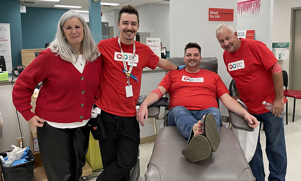 Man relaxing in donor bed after blood donation with his partner and partner’s parents at his side.