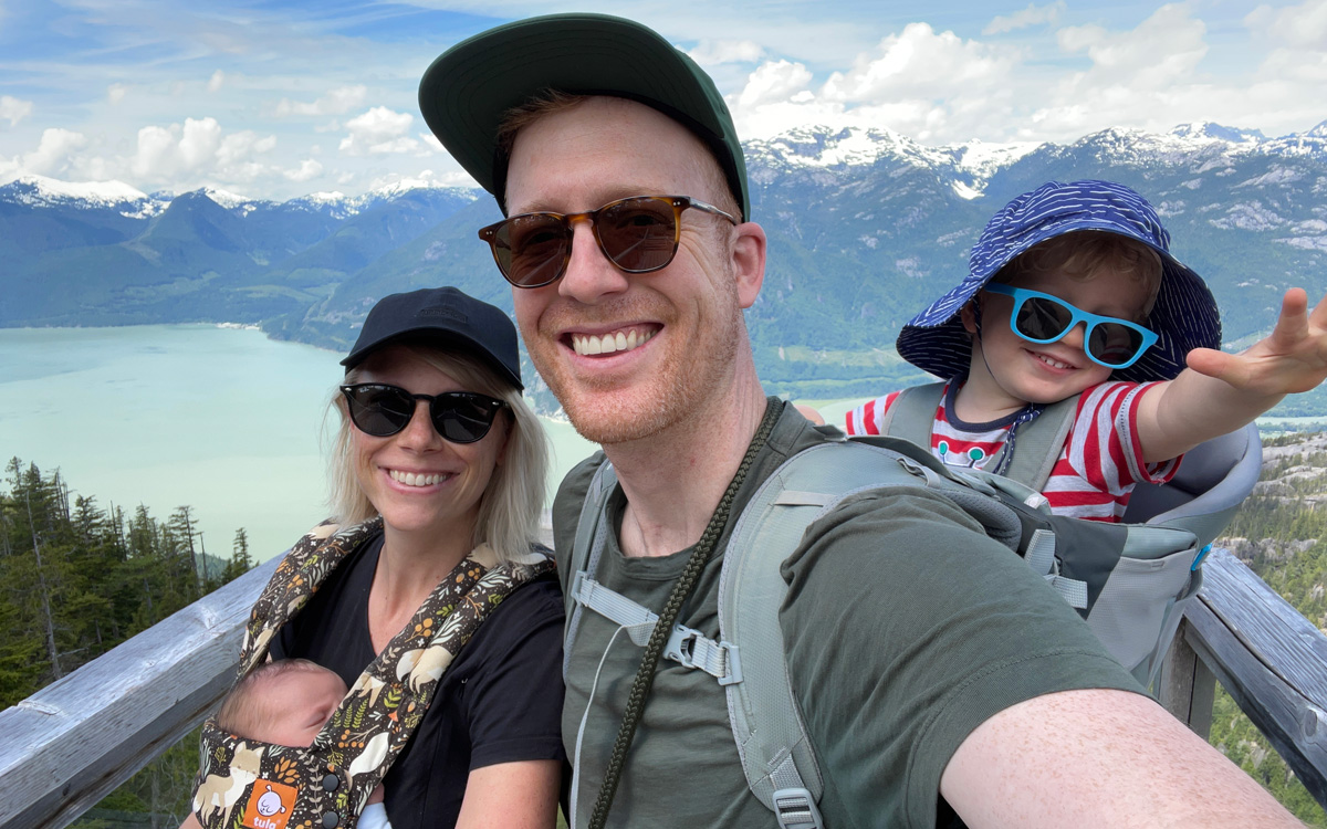 Dressed in casual outfits, Tyler and Rebecca Munk hang out with their toddler and baby on a bridge with surrounding mountains in Squamish, B.C.