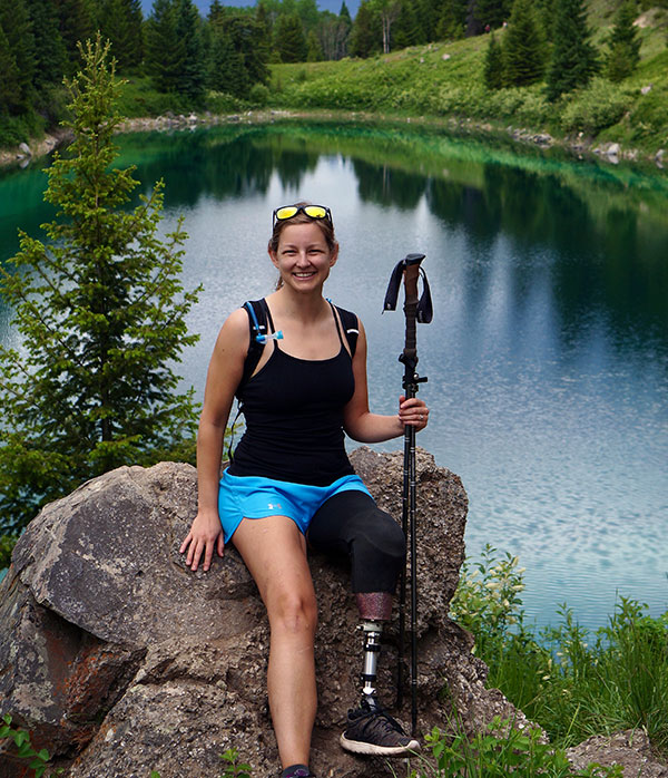 Woman with prosthetic left leg and walking poles sitting on a rock with lake and mountains in the background