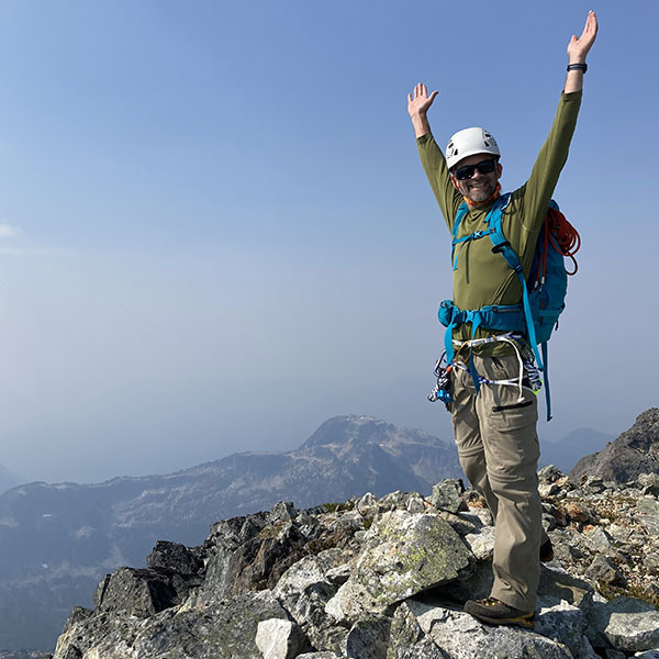 Man in mountain climbing gear raising hands atop Mount Doogie Dowler