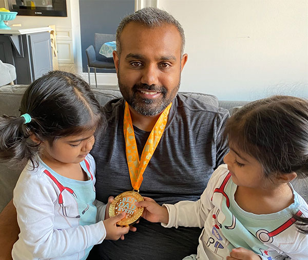Des jumelles tiennent une médaille accrochée autour du cou de leur père sur laquelle il est inscrit « Best Dad Ever » (meilleur papa du monde)