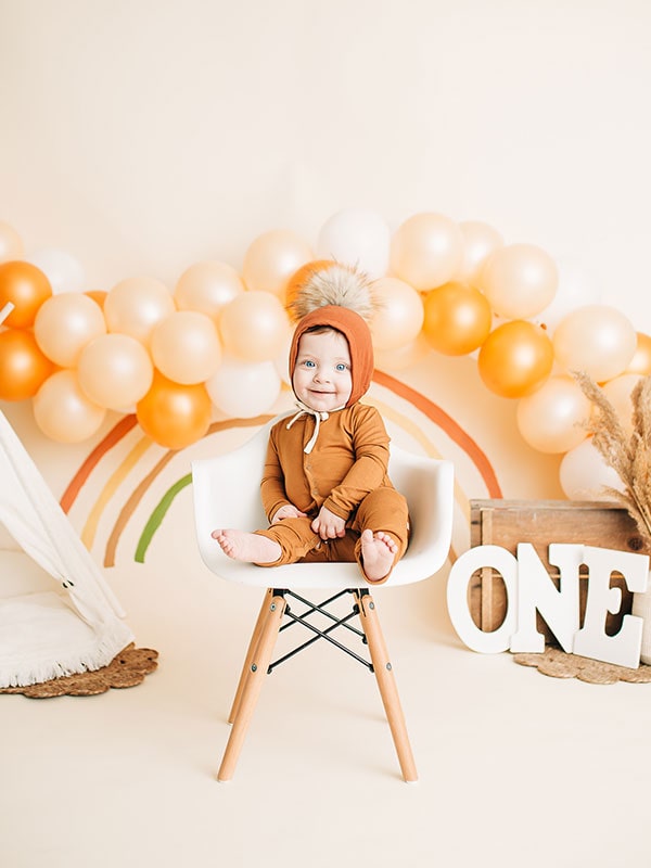Baby on chair in front of orange balloons and painted rainbow