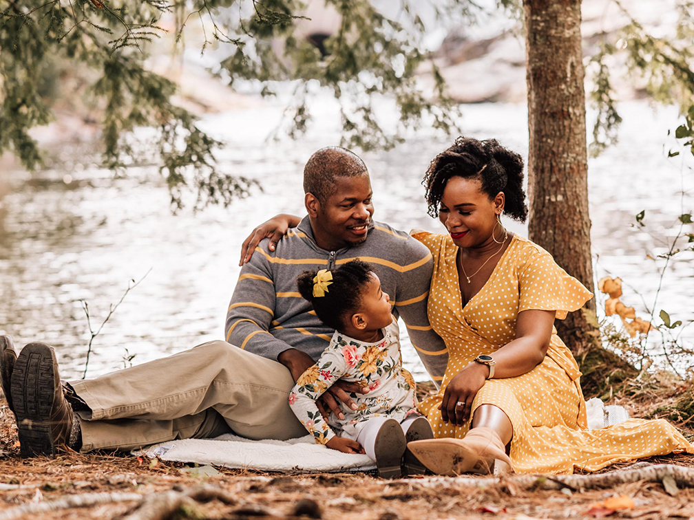 A family with a father, mother and young daughter, sit on a blanket among trees with water in the background.