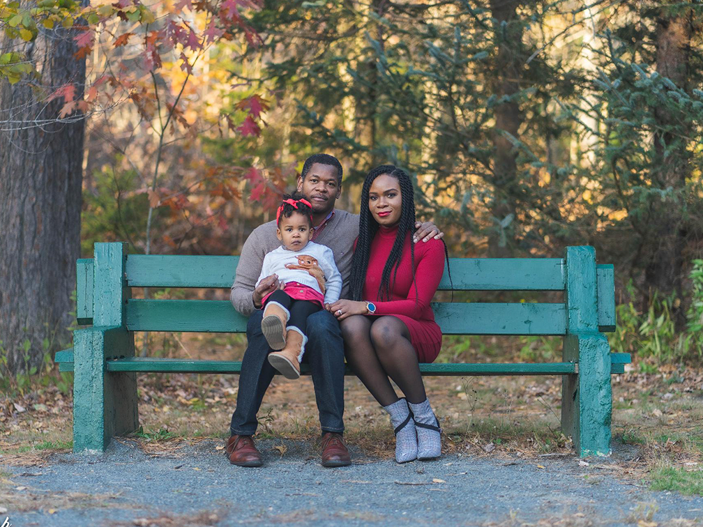 A family with a father, mother and young daughter, sit together on a park bench with a forest of large trees behind them.