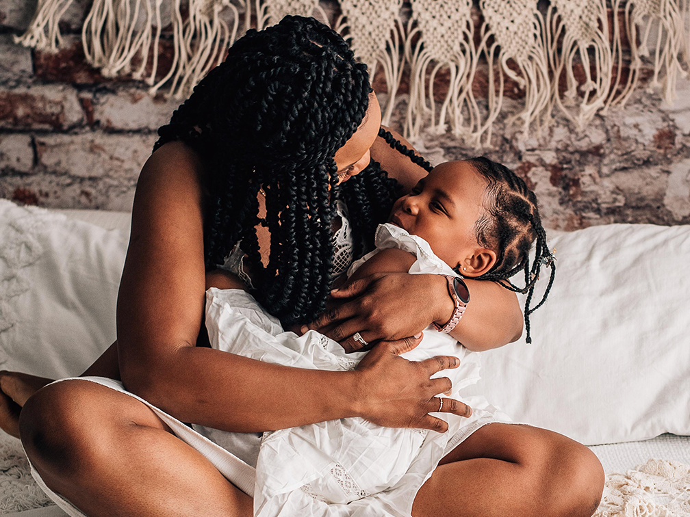 A mother gathers her young daughter into a full body embrace while sitting on a bed in front of a brick wall.