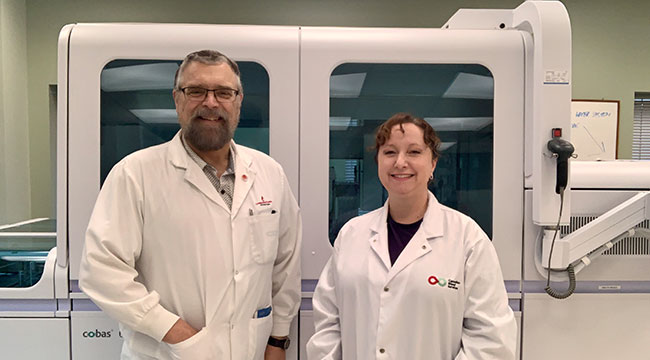 Douglas Ems & Valerie Conrod in front of a large machine wearing white lab coats