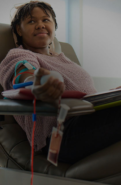 Visaree-mobile.jpg Darkly lit image of Visaree, sitting back in a donation chair while donating blood. Visaree is looking up at the nurse, while holding on to an exercise ball in the palm of her hand.