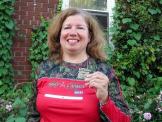 Image of Linda Paul holding her metal tags from the Army Run 5k event standing in front a house with bushes