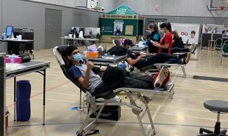 A donor gives a thumbs-up from the bed of a blood donation event hosted by the Ahmadiyya Muslim Youth Association. Other donors and Canadian Blood Services staff are in the background.