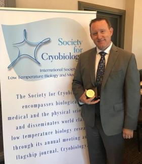 Dr Jason Acker holds his award in front of a banner for the Society for Cryobiology