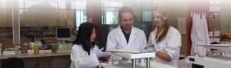 Image of 3 individuals wearing lab coats and gloves collaborating in a lab